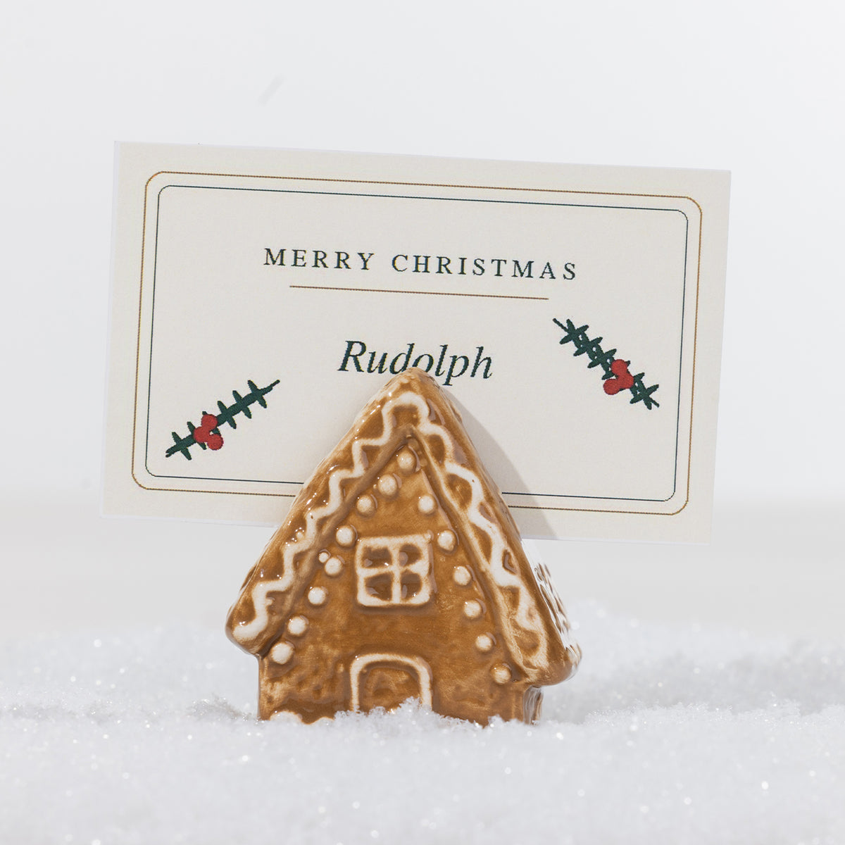 Gingerbread-style ceramic house name card holder with white icing details, holding a personalised Merry Christmas place card in a snowy setting.