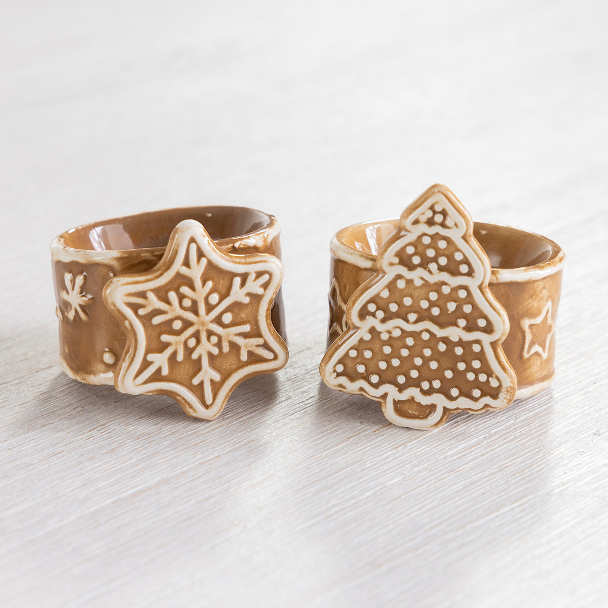 Gingerbread-style ceramic Christmas tree and snowflake napkin rings displayed together on a light timber surface.