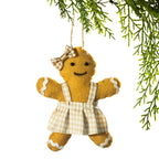 A gingerbread man felt Christmas ornament hanging from a green pine branch against a white background. The ornament is tan with a beige and white gingham dress and bow.