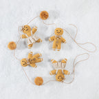 Four small felt gingerbread person ornaments in warm tan and cream tones, arranged on a white textured background with thin hanging strings and pom-pom accents.