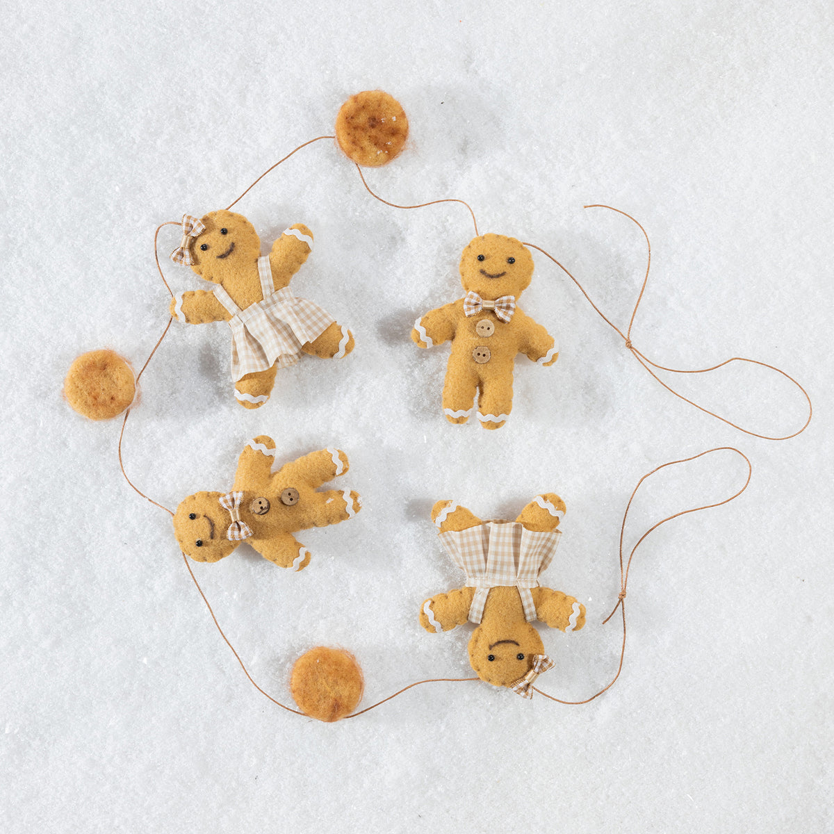 Four small felt gingerbread person ornaments in warm tan and cream tones, arranged on a white textured background with thin hanging strings and pom-pom accents.