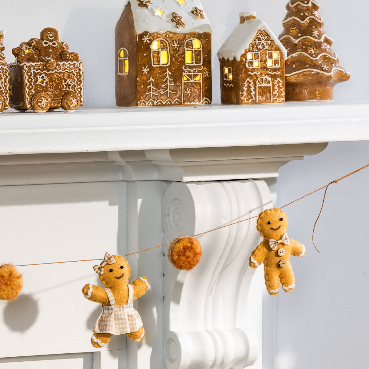 Gingerbread-themed holiday decor displayed on a white mantel, including gingerbread houses, a gingerbread tree, and a string garland with plush gingerbread figures and pom-poms.