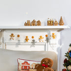 A cozy holiday scene with gingerbread-themed decor displayed on a white fireplace mantel and above a chair. The mantel holds small gingerbread houses and a tree, while a string of gingerbread men garlands hangs across the fireplace.