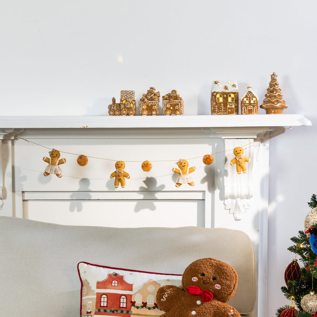 A cozy holiday scene with gingerbread-themed decor displayed on a white fireplace mantel and above a chair. The mantel holds small gingerbread houses and a tree, while a string of gingerbread men garlands hangs across the fireplace.