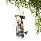 A small decorative hanging ornament shaped like a cow dressed as a chef, with a white hat, black-and-white plaid apron, and a small wooden spoon, suspended against a white background with green pine branches at the top.