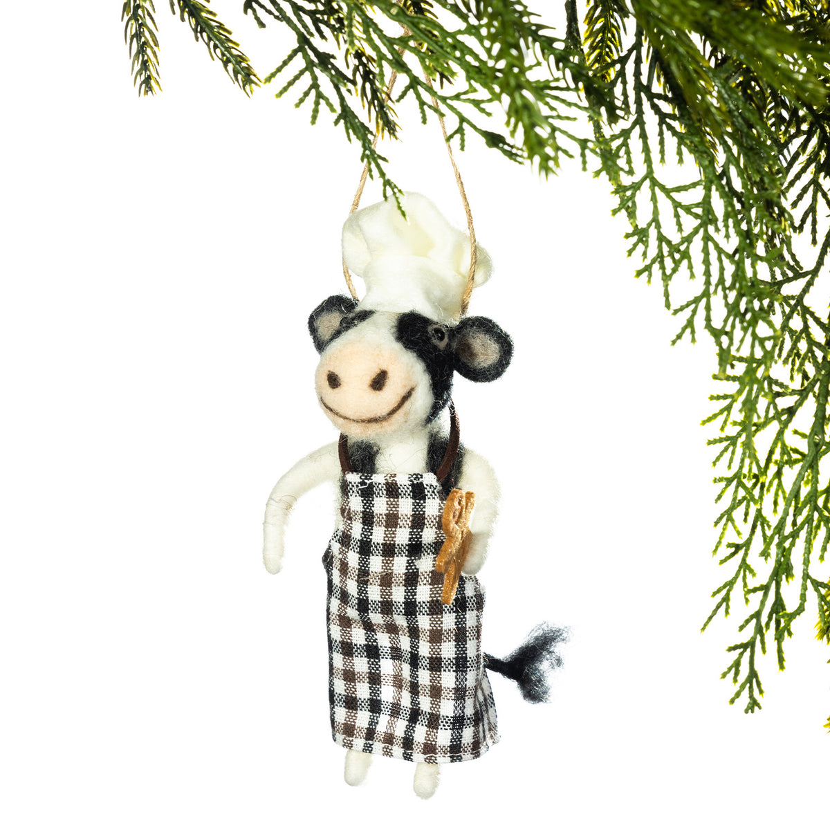 A small decorative hanging ornament shaped like a cow dressed as a chef, with a white hat, black-and-white plaid apron, and a small wooden spoon, suspended against a white background with green pine branches at the top.