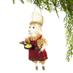 A felt sheep Christmas ornament dressed as a chef, wearing a white hat and red apron while holding a small frying pan with a yellow egg, hanging from a pine branch against a white background.