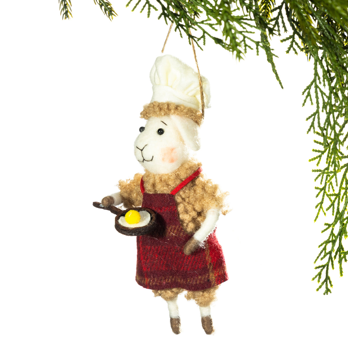 A felt sheep Christmas ornament dressed as a chef, wearing a white hat and red apron while holding a small frying pan with a yellow egg, hanging from a pine branch against a white background.