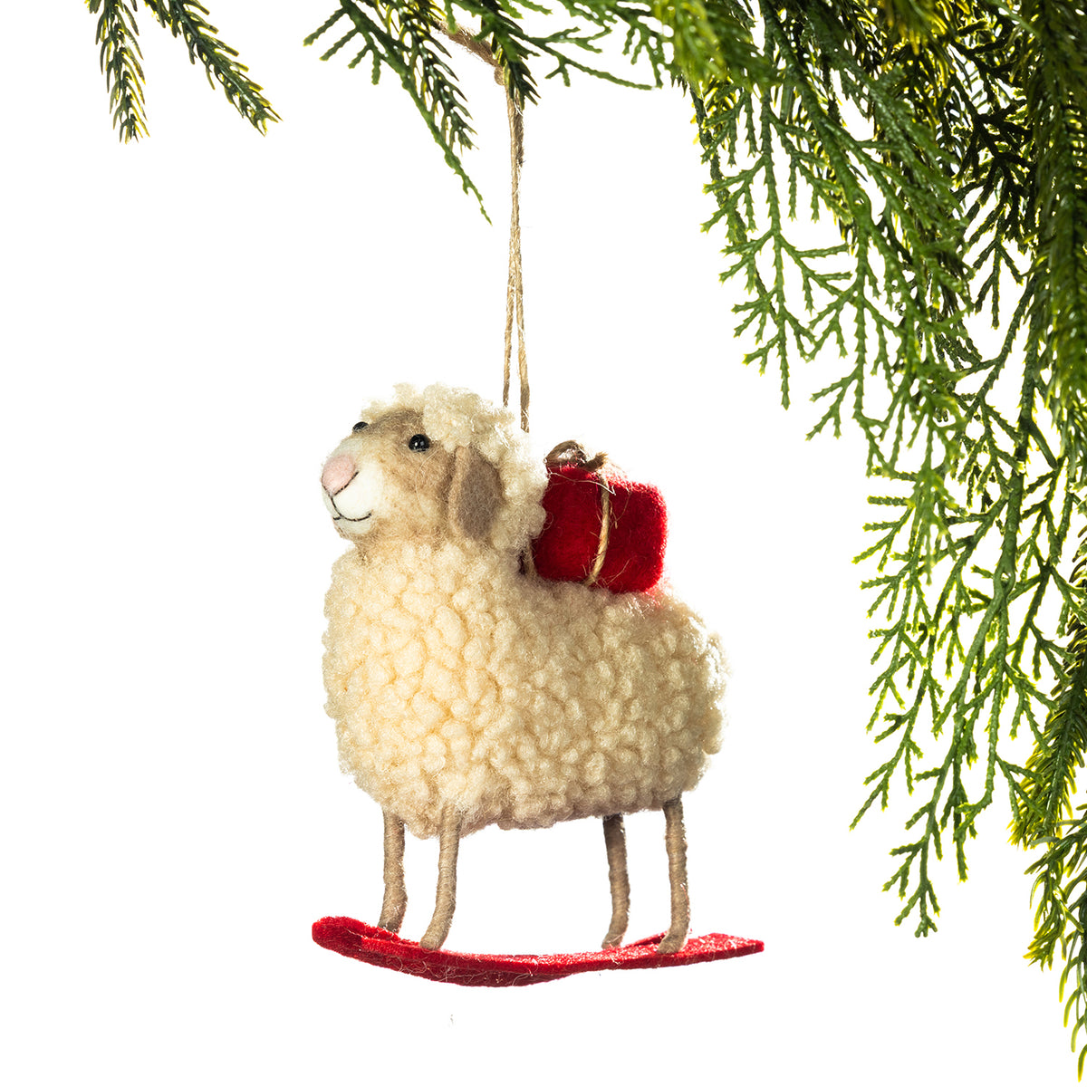 A felt sheep Christmas ornament with curly cream wool texture, a small red gift box on its back, and a red base, hanging from a tree branch against a white background.