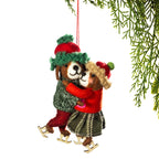 A felt Christmas ornament depicting two hugging dogs ice skating in winter clothing, hanging from a red string against a white background with pine branches at the top right.