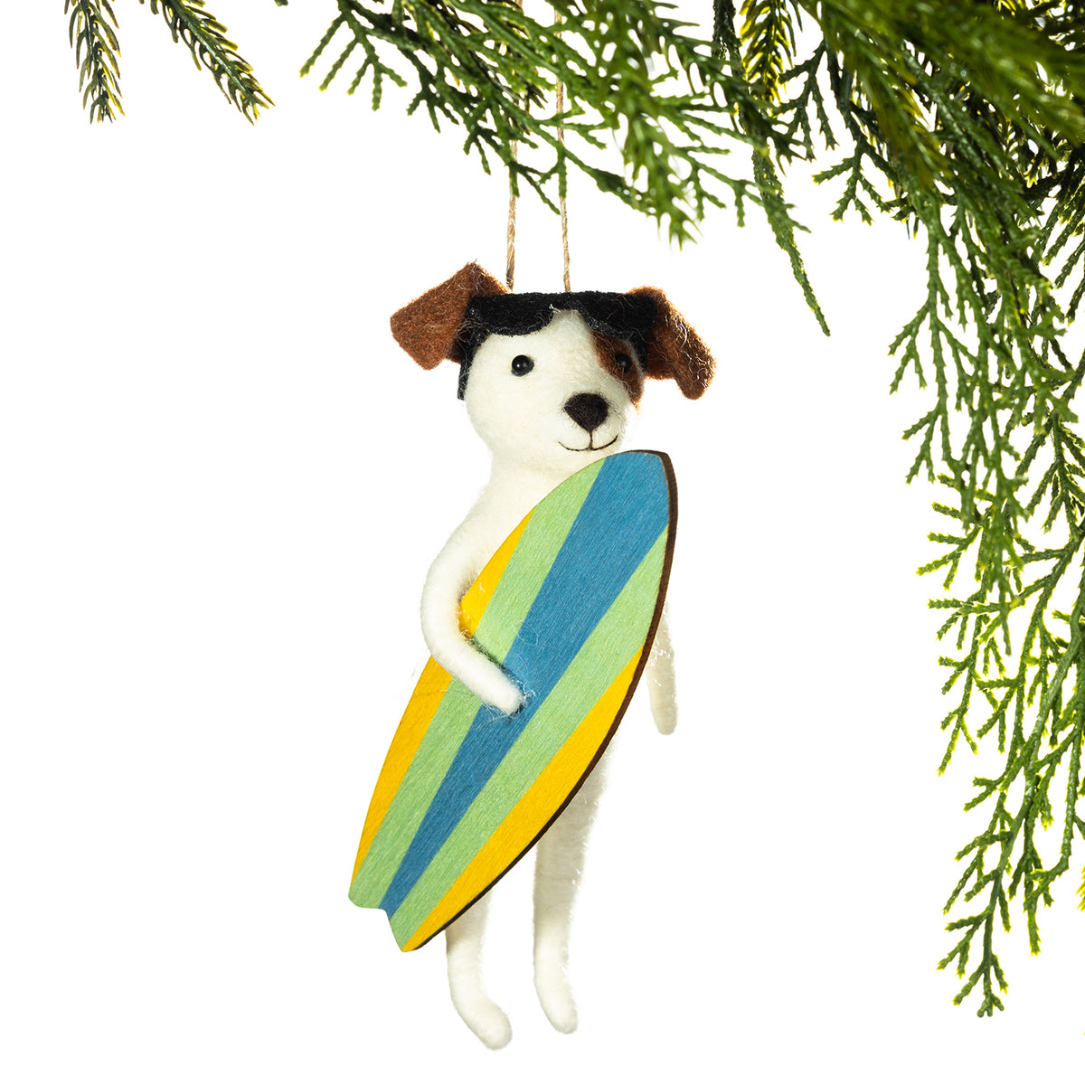 A felt dog Christmas ornament holding a striped surfboard in blue, green, yellow, and orange, hanging from a pine branch against a white background.