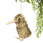 A small kiwi bird hanging ornament made of shaggy brown faux fur, with a long beak, small black eye, and tiny feet, suspended by a string against a white background with green pine branches at the top right.