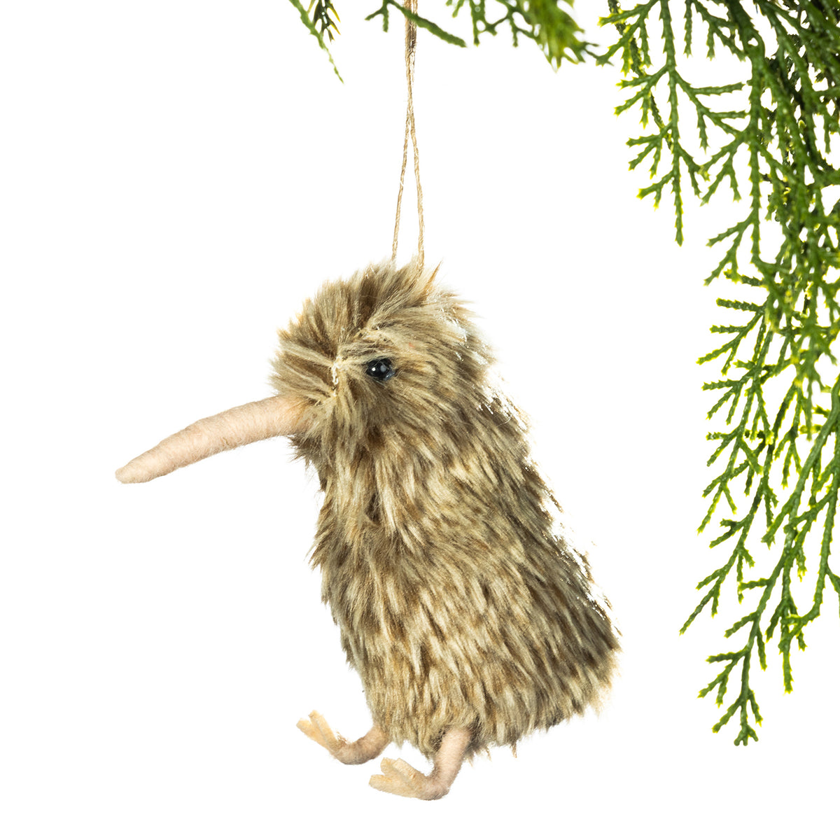 A small kiwi bird hanging ornament made of shaggy brown faux fur, with a long beak, small black eye, and tiny feet, suspended by a string against a white background with green pine branches at the top right.