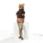 A knitted bear doll wearing a brown sweater and striped brown-and-beige socks, sitting on a white block against a white background with a snowy surface.