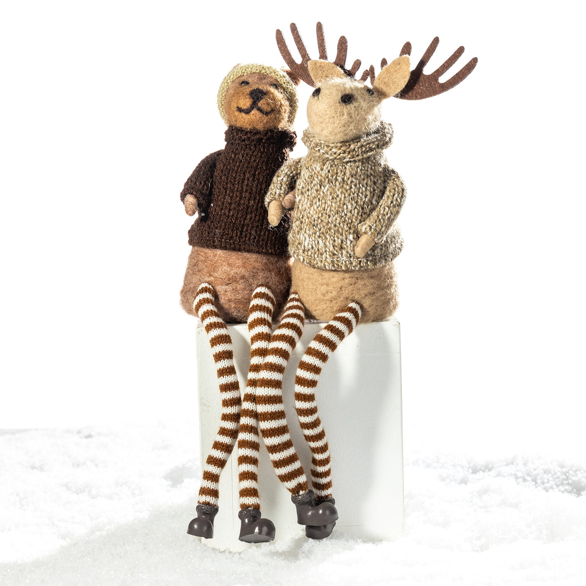 Two knitted moose figurines sitting side by side on a white block against a white background, one in dark brown and the other in beige, both wearing striped knit legs and winter clothing.