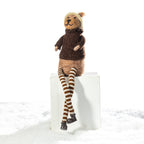 A knitted teddy bear doll wearing a brown sweater, tan pants, and striped brown-and-beige tights, sitting on a white block against a white background with a snowy surface.