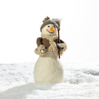 A small decorative snowman figurine standing in white faux snow, wearing a brown knit hat, cream scarf, and holding a twig staff, against a bright white background.