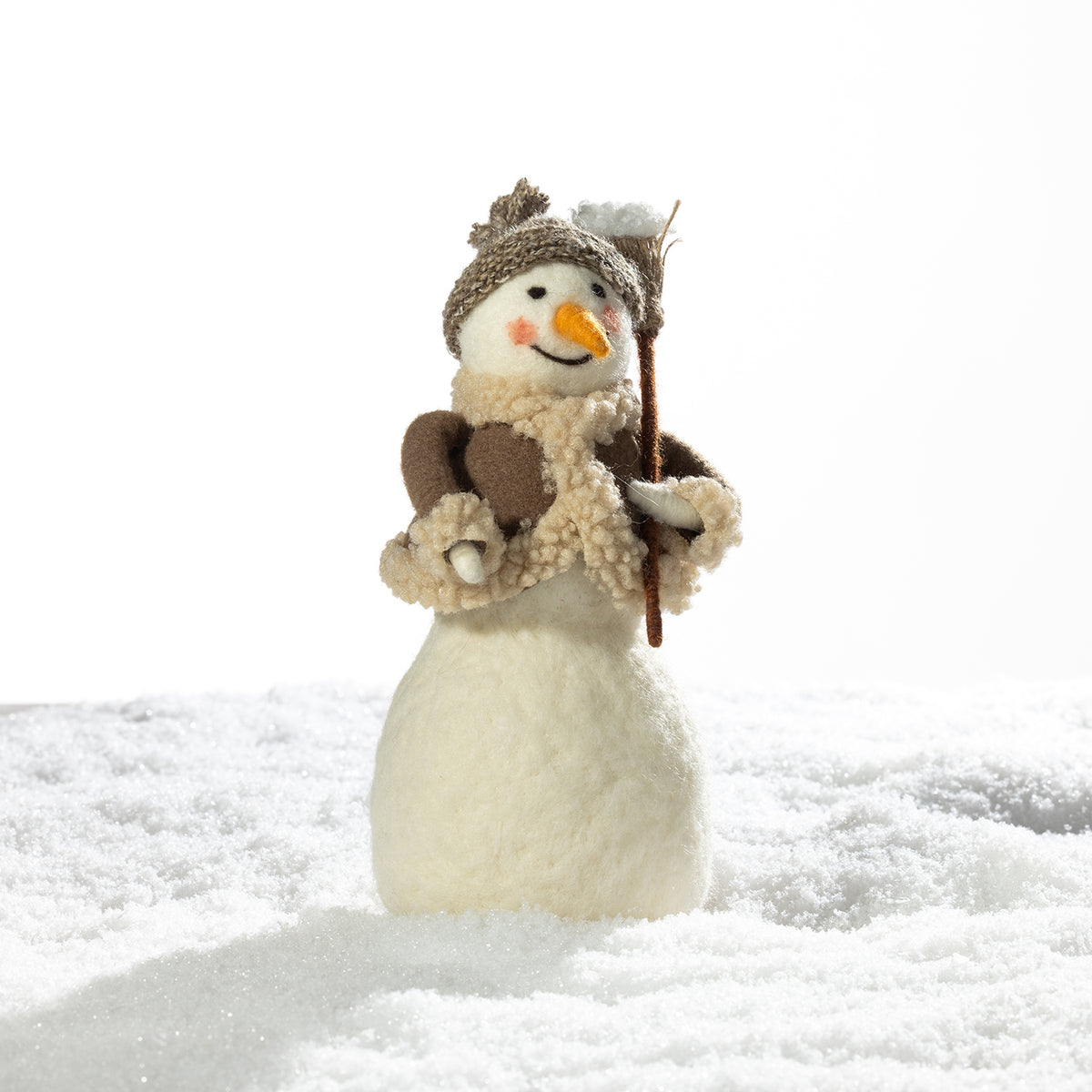 A small decorative snowman figurine standing in white faux snow, wearing a brown knit hat, cream scarf, and holding a twig staff, against a bright white background.