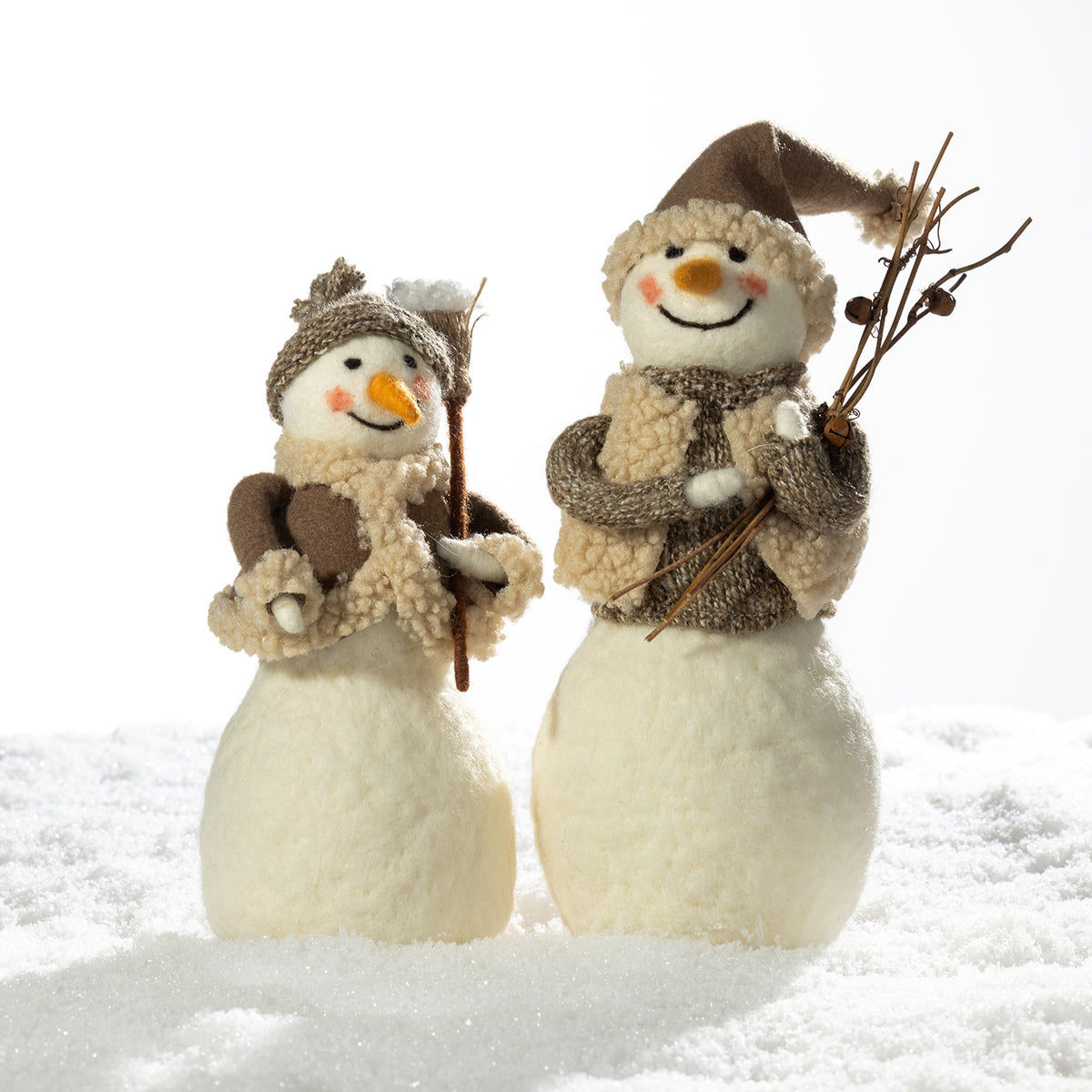 Two handmade felt snowman decorations standing in white faux snow against a bright white background. One smaller snowman wears a brown knit hat and scarf with a carrot nose and holds a twig.