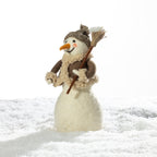 A small plush snowman figurine standing in white faux snow, wearing a brown knit hat, cream scarf, and holding a broom, photographed against a bright white background.