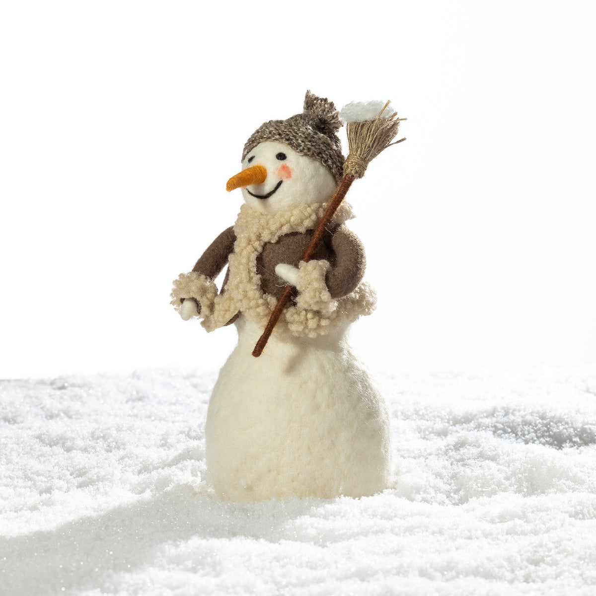 A small plush snowman figurine standing in white faux snow, wearing a brown knit hat, cream scarf, and holding a broom, photographed against a bright white background.