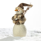 A felt snowman decoration standing in white faux snow against a bright white background. The snowman has a brown pointed hat, a cream textured body, a brown scarf, and holds small twig branches.