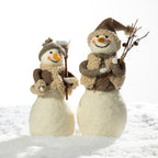 Two handmade felt snowman decorations standing in white faux snow against a bright white background. One smaller snowman wears a brown knit hat and scarf with a carrot nose and holds a twig.