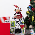 A plush cow doll dressed in a red Santa hat, red plaid skirt, and red scarf, sitting on Christmas-themed gift boxes next to a decorated Christmas tree.