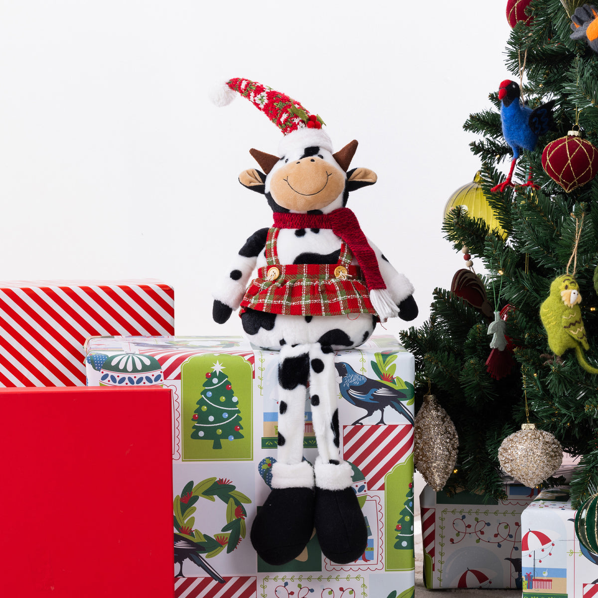 A plush cow doll dressed in a red Santa hat, red plaid skirt, and red scarf, sitting on Christmas-themed gift boxes next to a decorated Christmas tree.