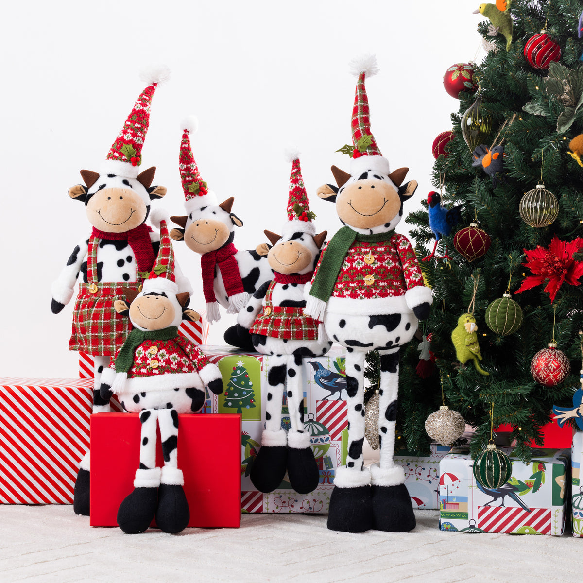 A set of five Christmas cow plush figurines in different sizes, wearing festive red and green holiday sweaters, plaid skirts, and pointed red hats, posed next to a decorated Christmas tree and wrapped gifts.