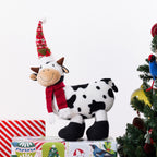 A plush cow figurine with black and white spots, wearing a red knit scarf and a red Christmas hat, standing on top of wrapped gifts next to a decorated Christmas tree.