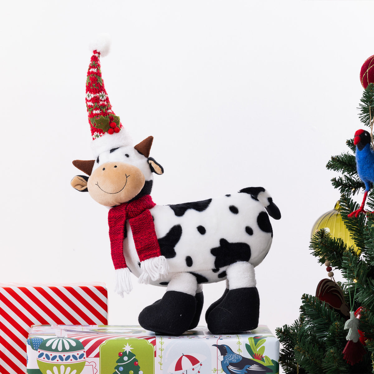 A plush cow figurine with black and white spots, wearing a red knit scarf and a red Christmas hat, standing on top of wrapped gifts next to a decorated Christmas tree.
