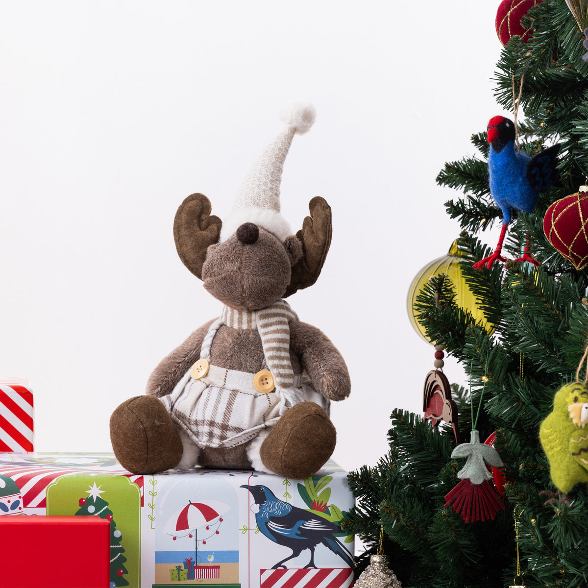 A plush moose figurine wearing a beige knit Santa hat, striped scarf, and plaid skirt, sitting on wrapped Christmas presents beside a decorated Christmas tree with a blue bird ornament.