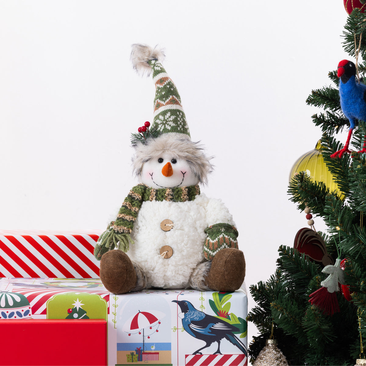 A plush snowman figurine wearing a knitted green and cream hat and scarf, sitting on wrapped Christmas presents beside a decorated Christmas tree.