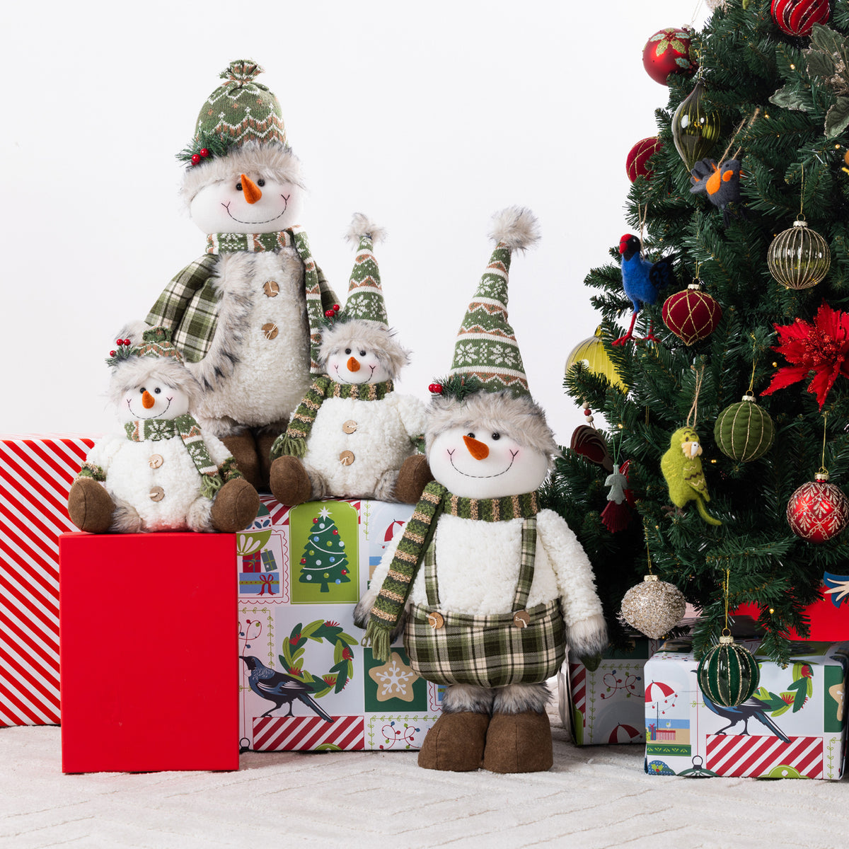 Plush snowmen ornaments in different sizes arranged on wrapped Christmas presents beside a decorated Christmas tree. The snowmen wear green plaid scarves and pointed green knit hats with white fur bodies and orange carrot noses.