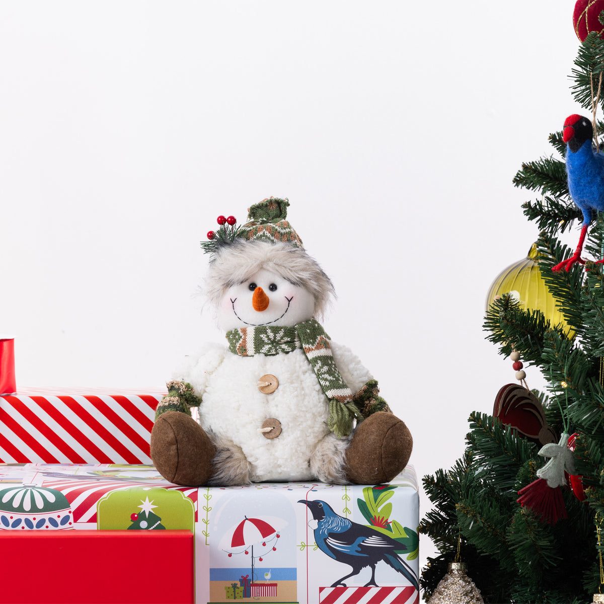 A plush snowman figurine sits on top of wrapped Christmas presents, with a decorated Christmas tree visible on the right against a plain white background.