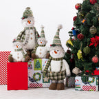 Plush snowmen figurines in different sizes arranged on wrapped Christmas presents beside a decorated Christmas tree. The snowmen wear green plaid scarves and pointed green knit hats with white fur bodies and orange carrot noses.