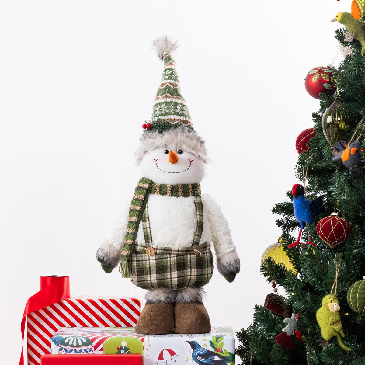 A plush snowman figurine wearing a green plaid hat and scarf stands on wrapped gifts beside a decorated Christmas tree against a white background.