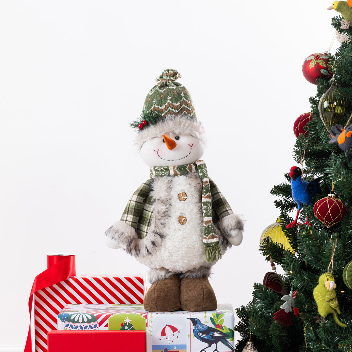 A plush Christmas snowman figurine stands on wrapped gifts beside a decorated Christmas tree. The snowman wears a green knit hat and scarf with brown boots and an orange carrot nose.