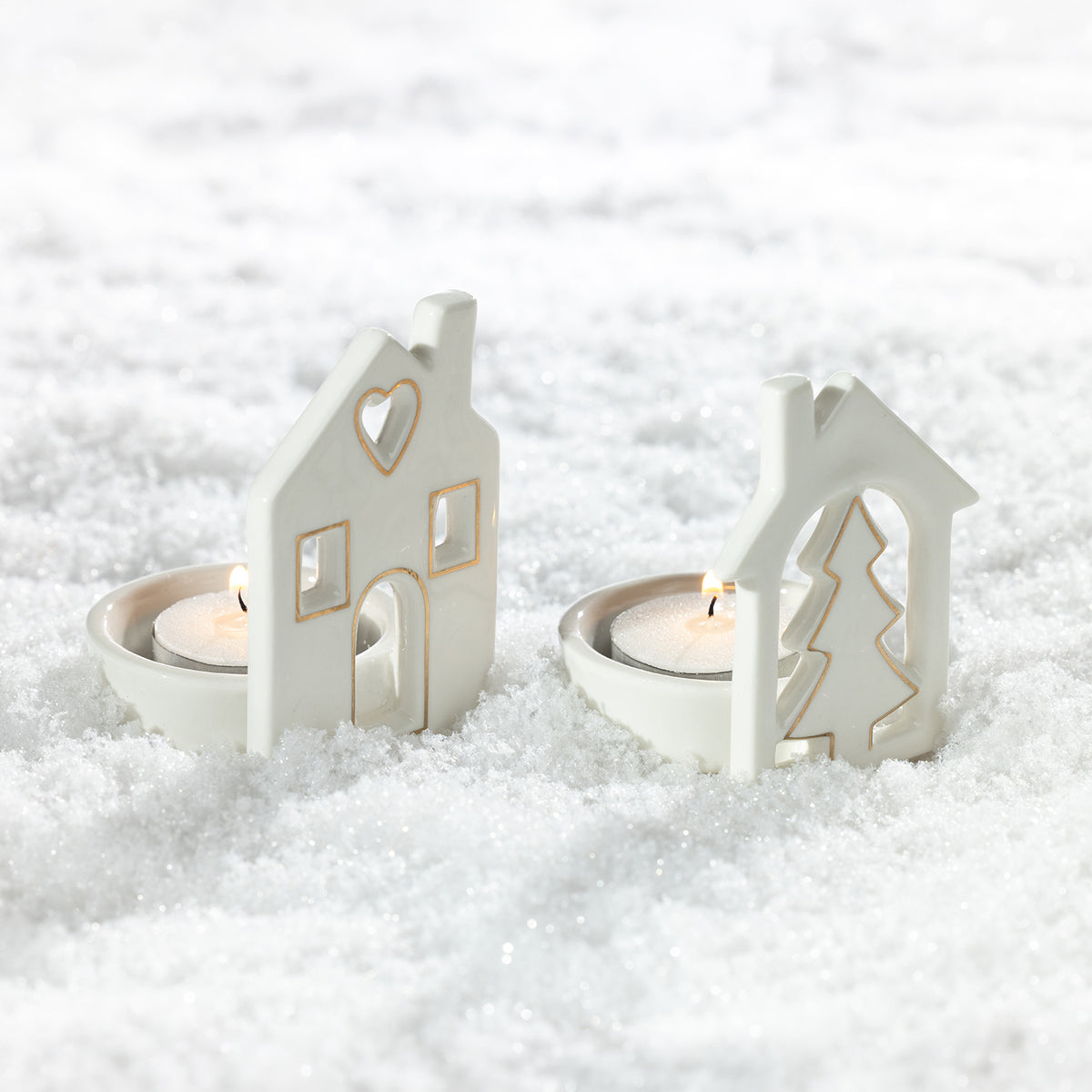 Two white ceramic Christmas house tealight candle holders with gold detailing, featuring heart, window and Christmas tree designs, displayed in artificial snow with lit candles.