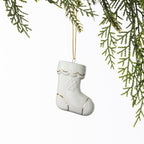 White ceramic Christmas stocking hanging decoration with delicate gold detailing and an embossed snowflake design, suspended from a gold string amongst green foliage.
