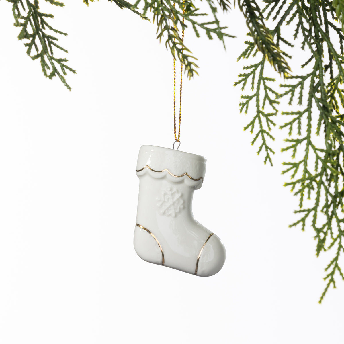 White ceramic Christmas stocking hanging decoration with delicate gold detailing and an embossed snowflake design, suspended from a gold string amongst green foliage.