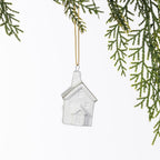 White ceramic Christmas house hanging decoration with embossed windows, a doorway and delicate gold roof detailing, suspended from a gold string amongst green foliage.