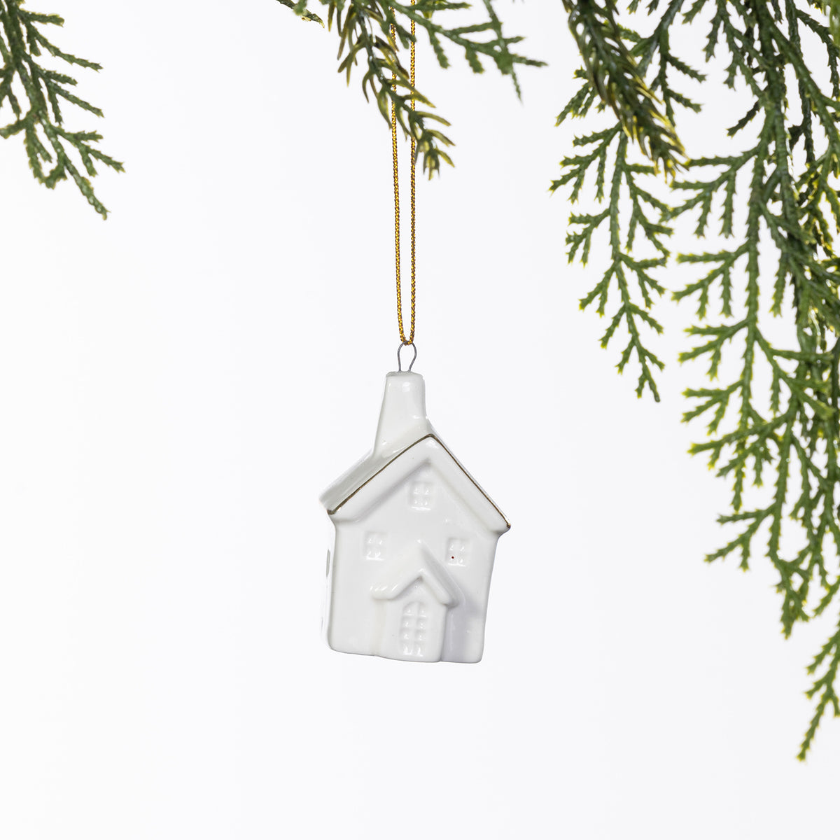 White ceramic Christmas house hanging decoration with embossed windows, a doorway and delicate gold roof detailing, suspended from a gold string amongst green foliage.