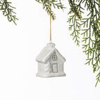 White ceramic Christmas house hanging decoration with a textured snow-covered roof and delicate gold window and door detailing, suspended from a gold string amongst green foliage.