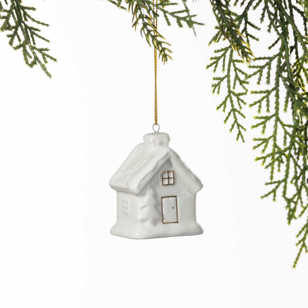 White ceramic Christmas house hanging decoration with a textured snow-covered roof and delicate gold window and door detailing, suspended from a gold string amongst green foliage.