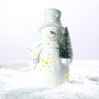 White ceramic snowman LED decoration wearing a top hat and scarf, holding a miniature Christmas tree with glowing star and dot cut-outs on a snowy surface.