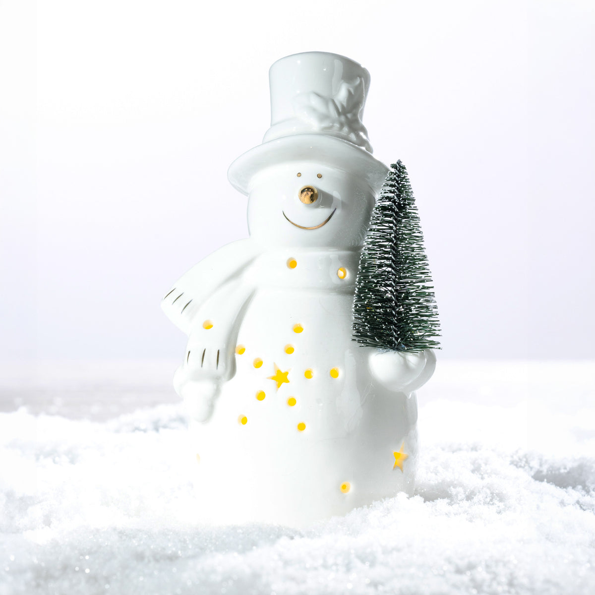White ceramic snowman LED decoration wearing a top hat and scarf, holding a miniature Christmas tree with glowing star and dot cut-outs on a snowy surface.