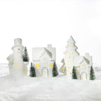 A festive display of illuminated white ceramic Christmas houses, a snowman and a Christmas tree surrounded by miniature green trees on a snowy surface.