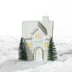 Small white ceramic brick house LED decoration featuring glowing windows, gold-trimmed details and two miniature Christmas trees on a snowy surface.